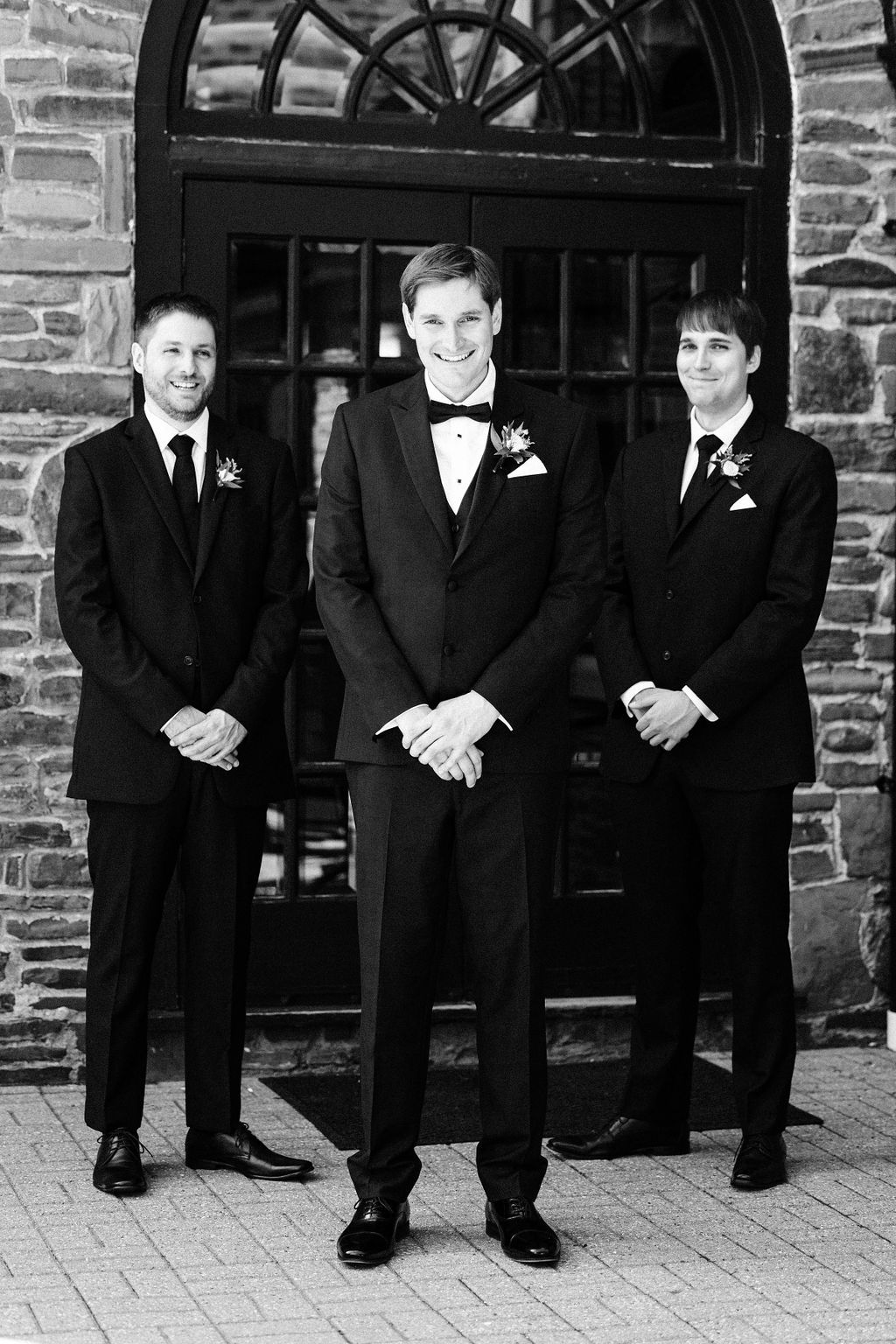 Groomsmen in black tuxedos stand together against a brick wall in a candid wedding photo series.