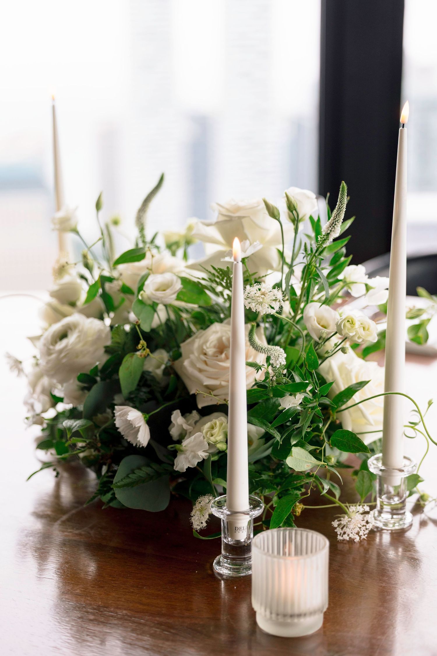 White floral centerpiece with taper candles and greenery on wooden dining table.