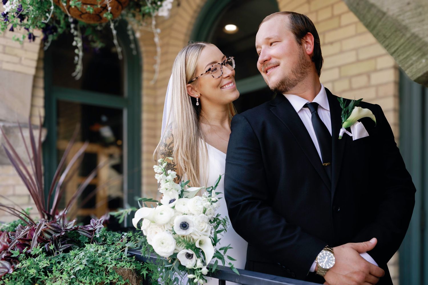 A couple in wedding attire share a romantic moment in front of a brick building with white flowers and greenery.