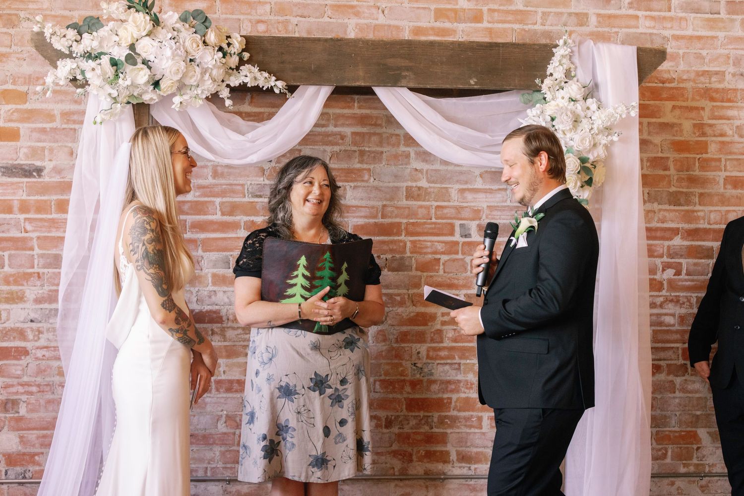 Elegant wedding ceremony setup with white floral arch and draped fabric against exposed brick wall.