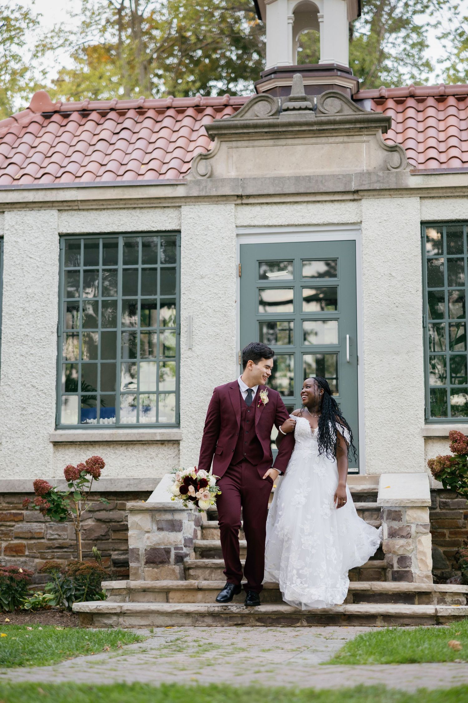 Bride in white lace gown and groom in burgundy suit hold hands on stone steps outside elegant building.