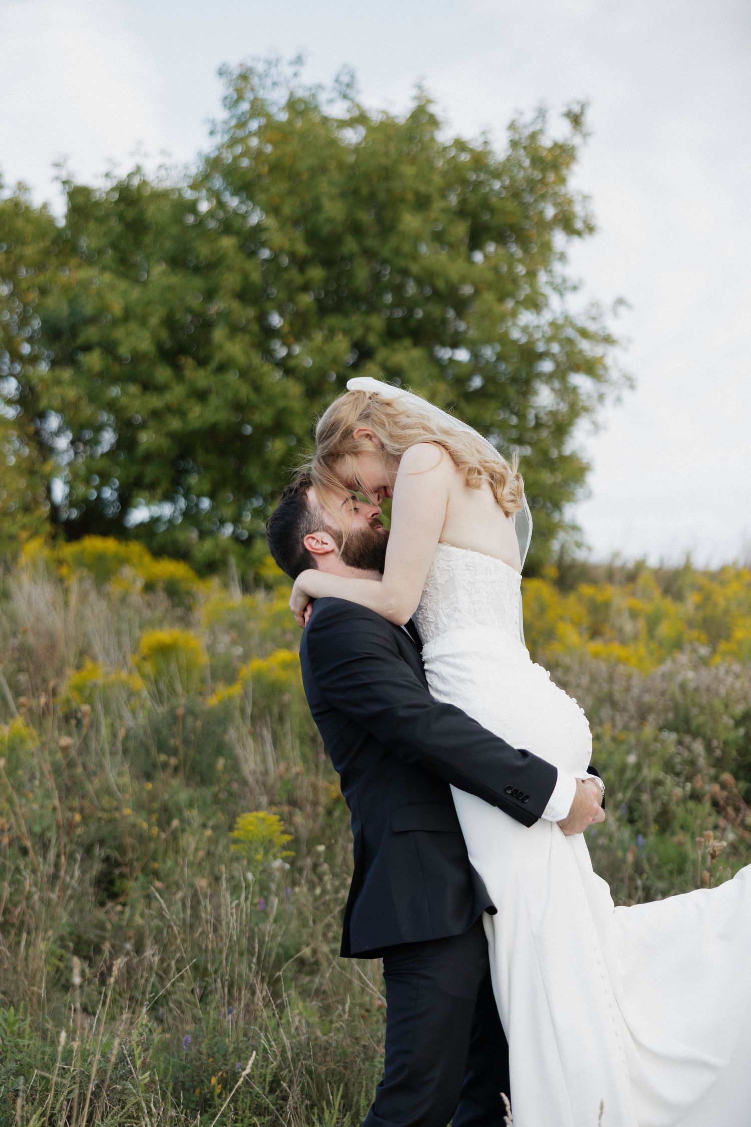 Groom lifting bride in white gown outdoors, sharing a kiss surrounded by wildflowers and green trees.