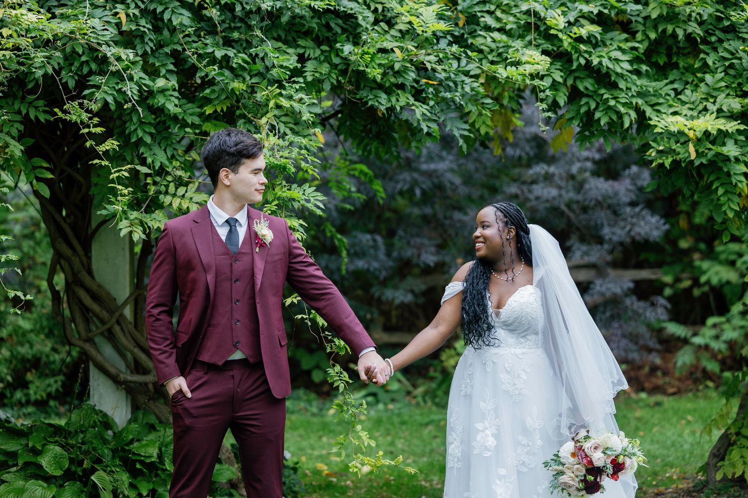 Interracial couple in wedding attire holding hands in lush garden, groom in burgundy suit, bride in white gown with veil.