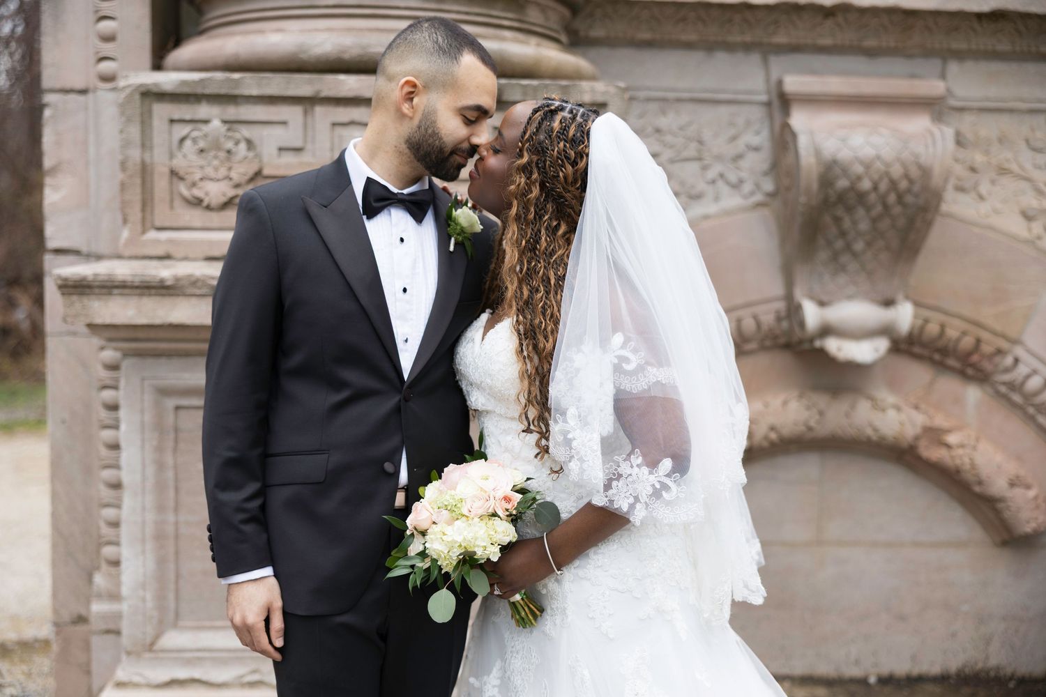 Interracial couple sharing a romantic moment in wedding attire, bride holding bouquet, groom in tuxedo, stone architecture backdrop.