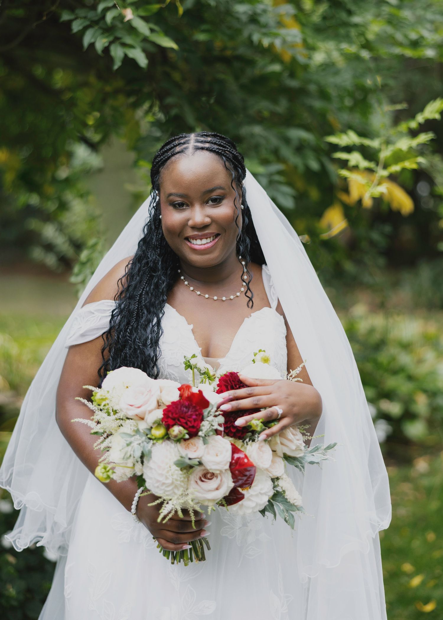 Bride in white wedding dress holding red and white bouquet against natural garden backdrop.