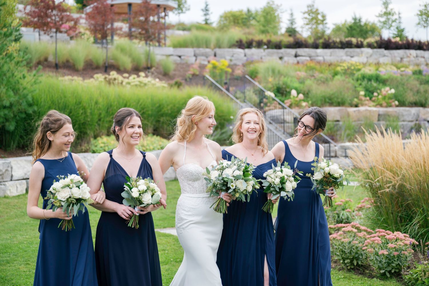 Bridesmaids in navy dresses and bride in white gown pose together in a lush garden setting during summer wedding.