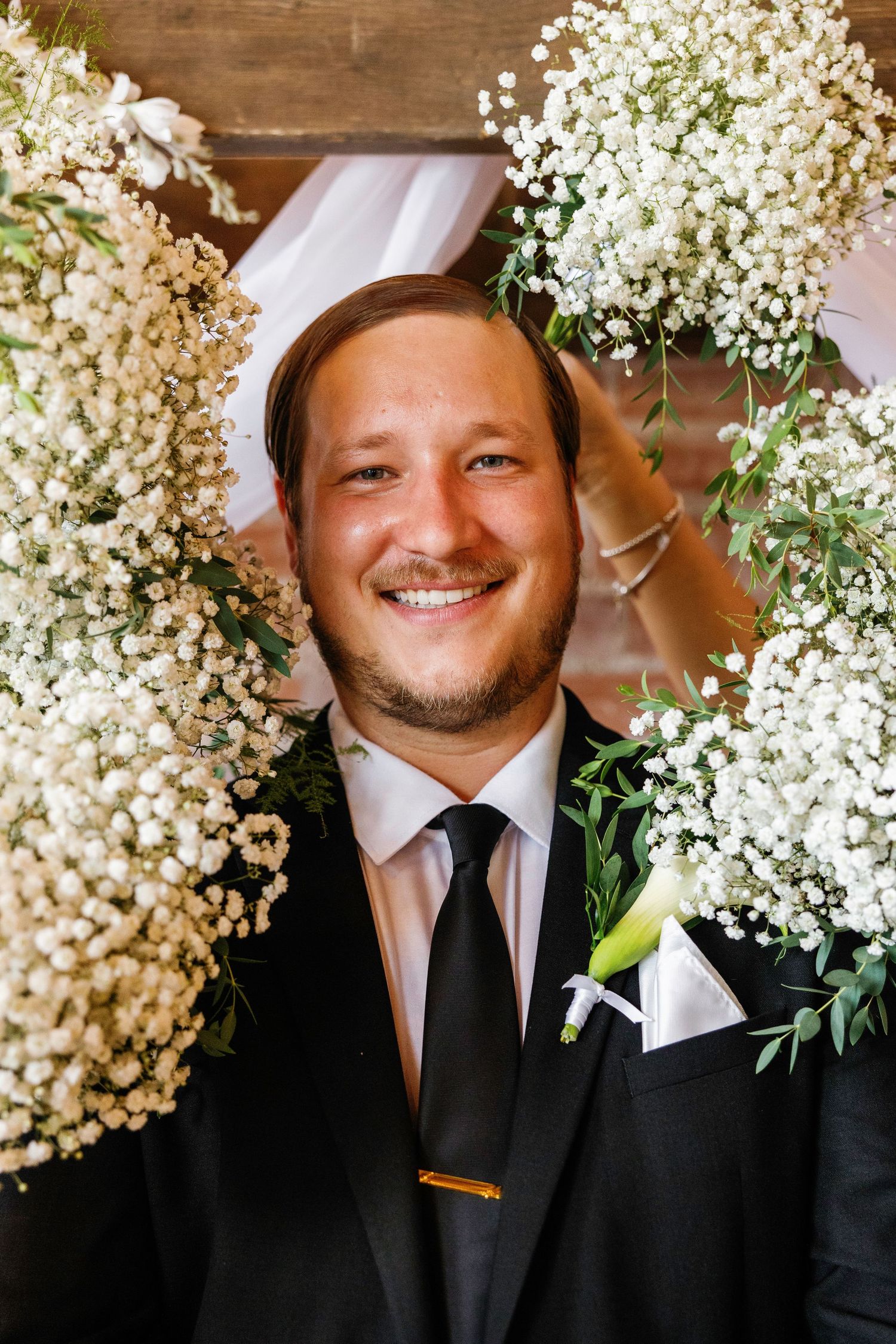 Groomsman in black suit and pink tie smiling warmly at wedding ceremony with white floral decorations.