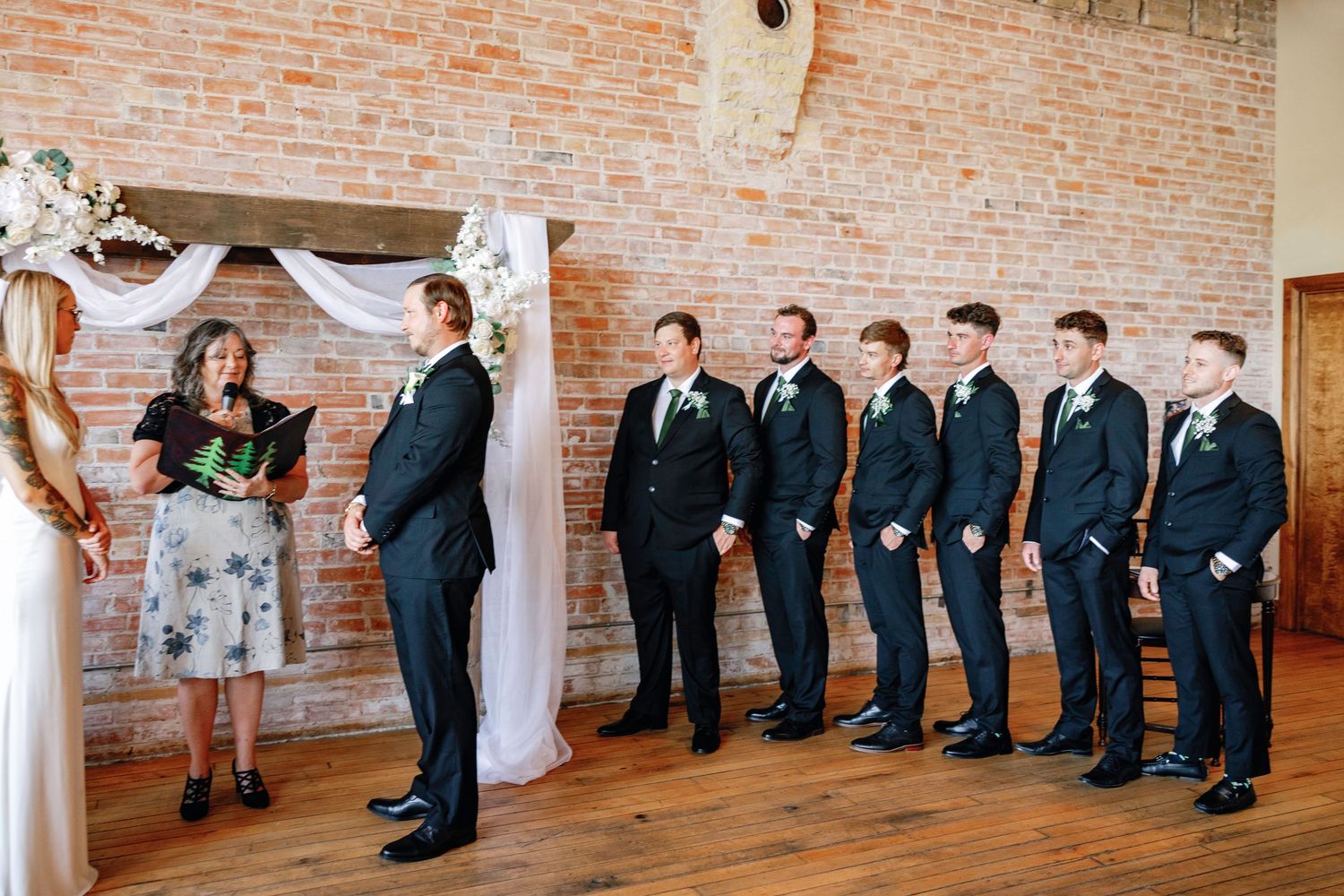 Wedding party lined up against exposed brick wall in rustic indoor venue with elegant white drapery.