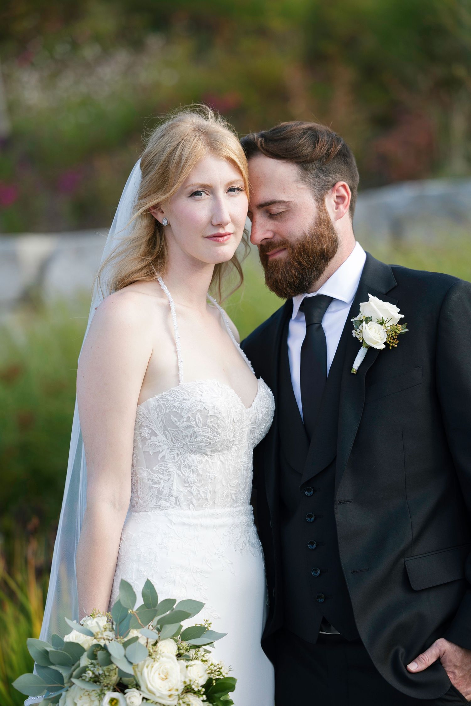 Bride and groom share a tender moment outdoors, bride in lace gown with veil holding white roses bouquet.