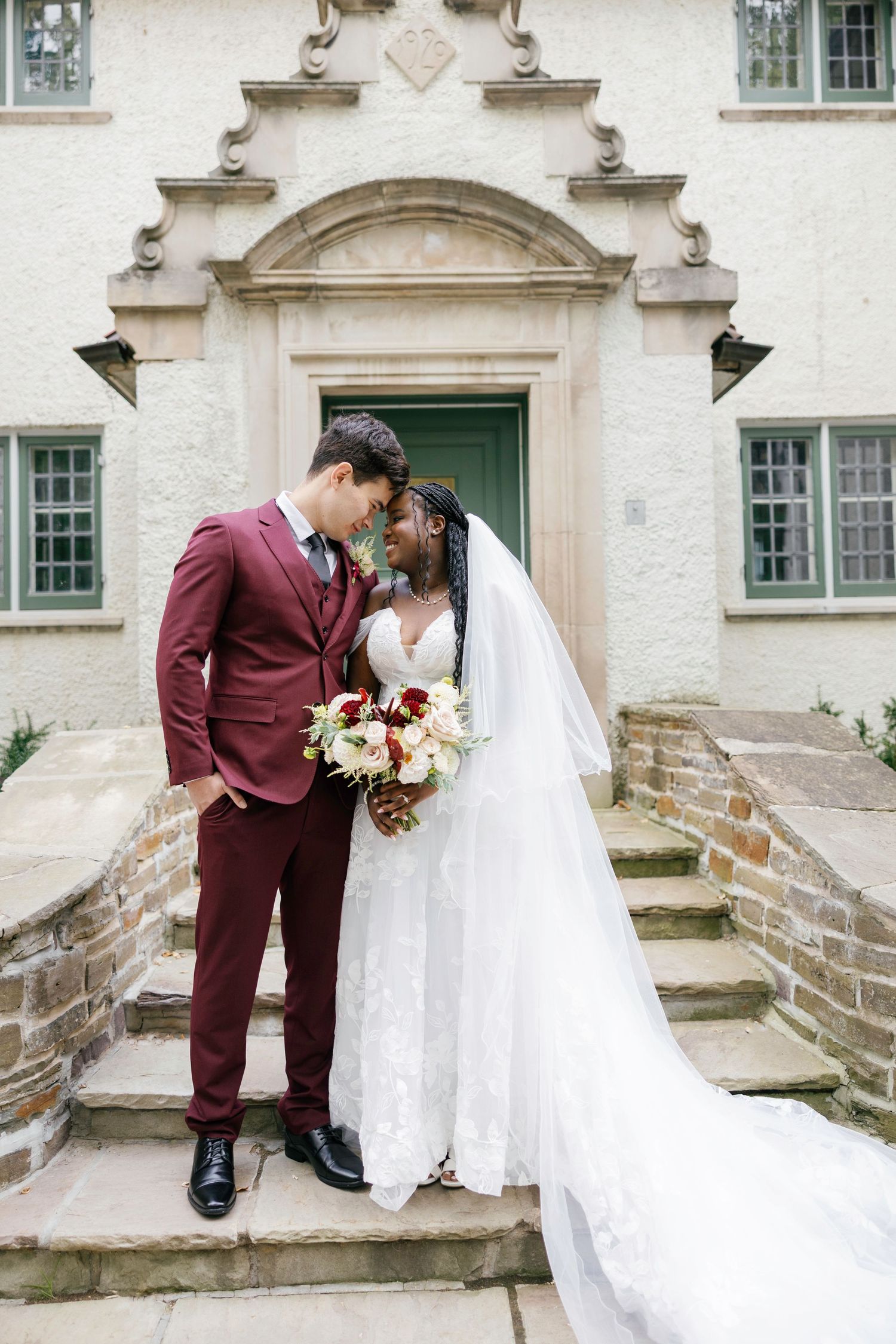 Bride and groom share a romantic moment on stone steps outside a historic venue, she in lace gown with veil, he in burgundy suit.