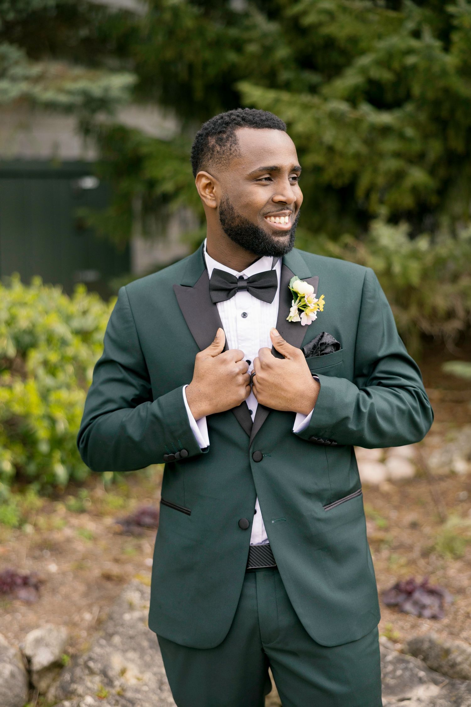 Smiling groom in green tuxedo with black bow tie and boutonniere posing outdoors at wedding.