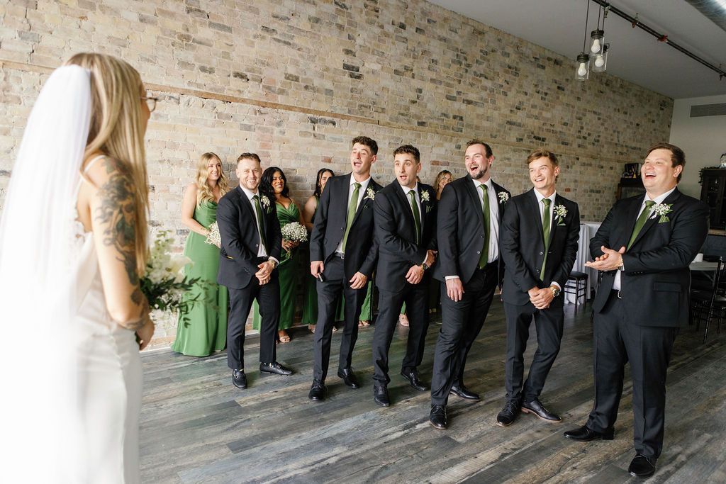 Groomsmen in black suits with green ties react joyfully as bride in white veil reveals her wedding look indoors.