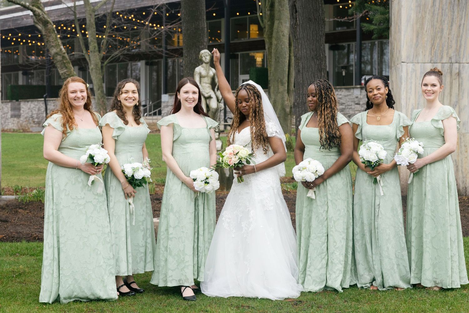Bride in white gown poses with five bridesmaids in sage green dresses holding white bouquets outdoors.