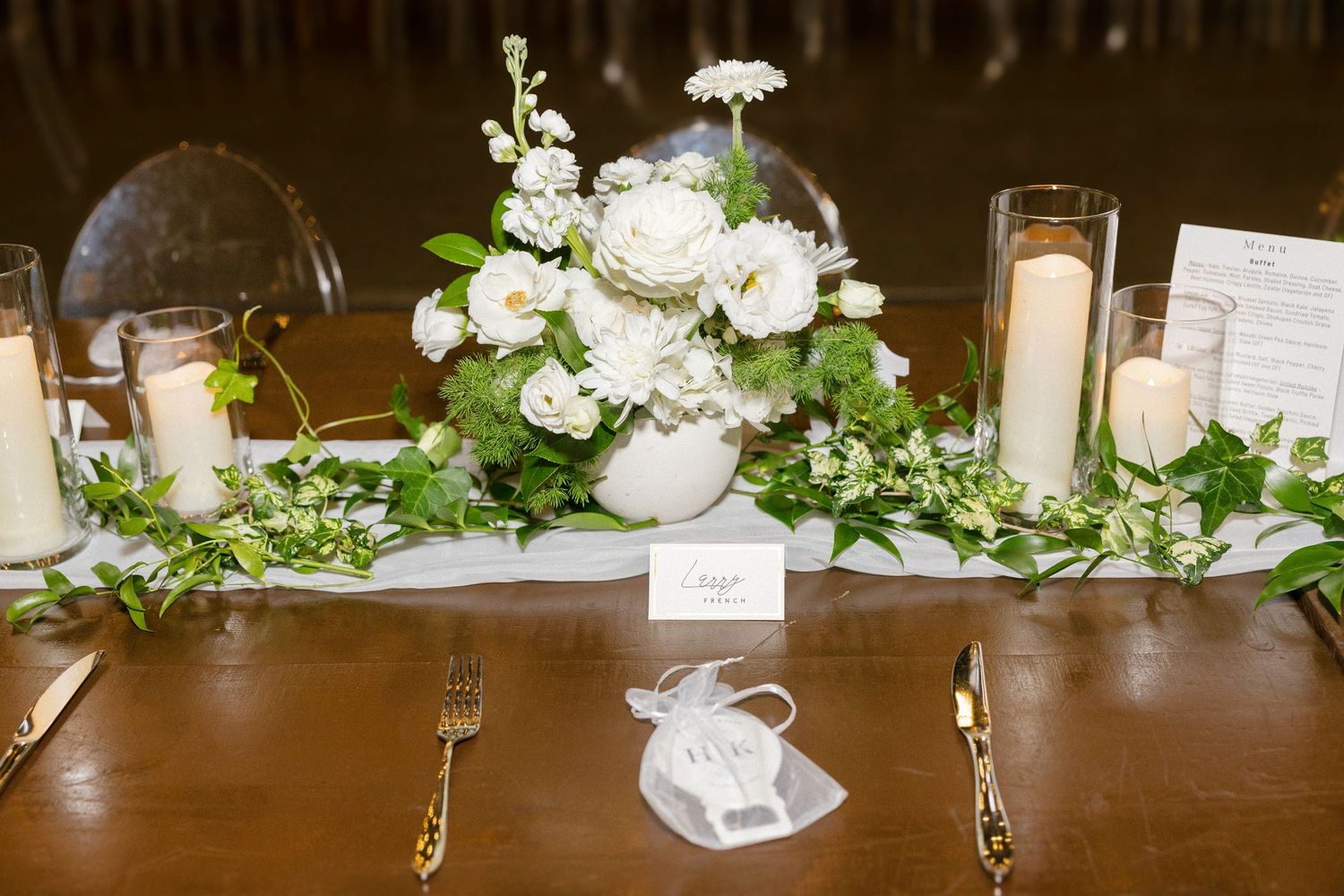 Elegant wedding table setting with white floral centerpiece, greenery garland, gold cutlery, and white candles.