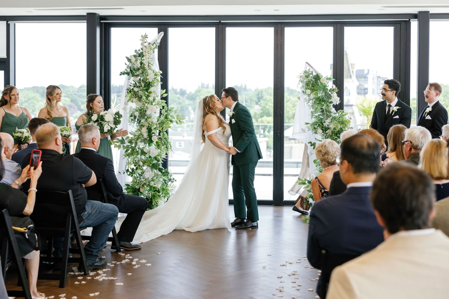 Indoor wedding ceremony with large windows, featuring a couple exchanging vows in front of seated guests.