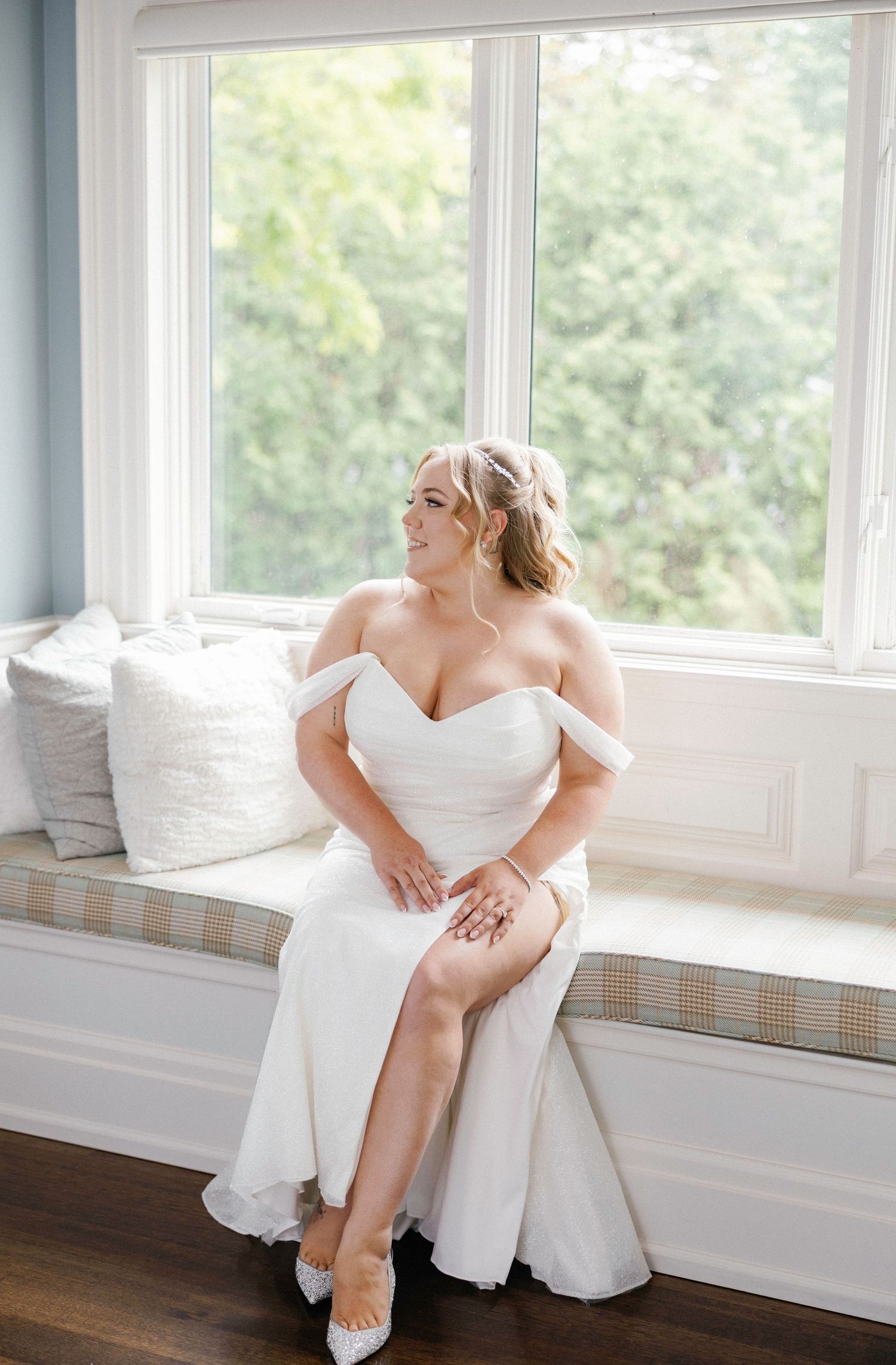 Bride sitting on window bench adjusts her shoe in natural lighting.