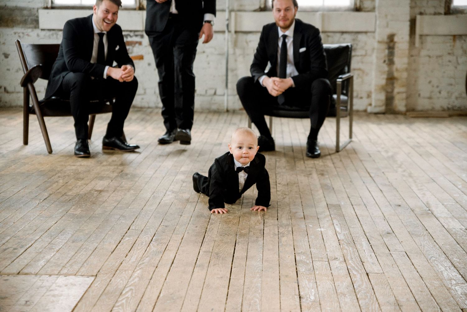Groomsmen in black suits sit casually on wooden floor of industrial space with white brick walls.