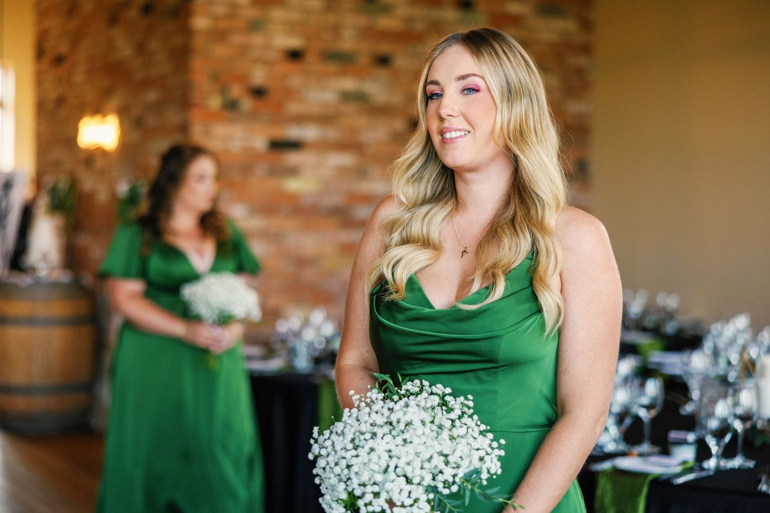 Bridesmaids in emerald green dresses hold white baby's breath bouquets in an elegant indoor venue.