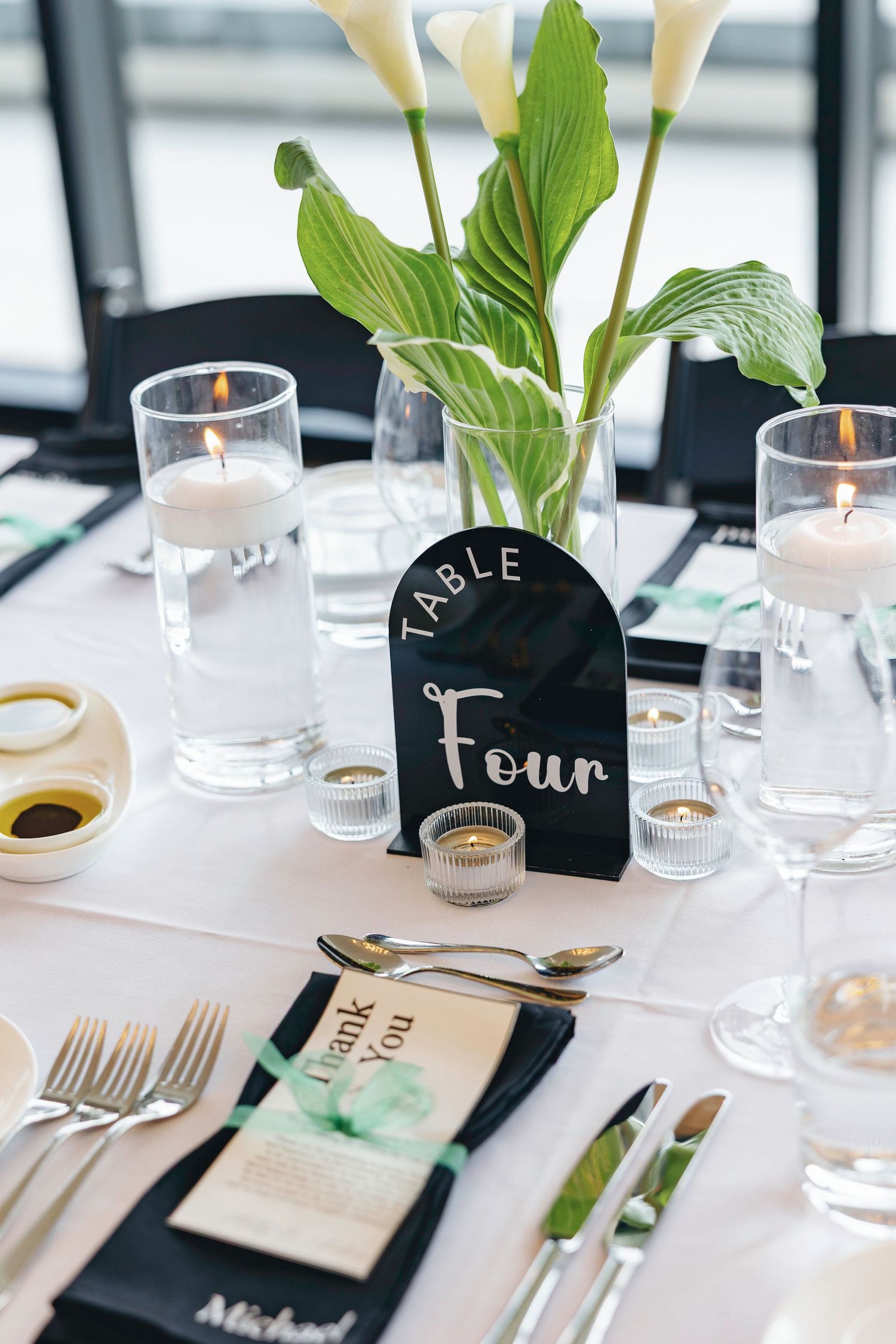 Elegant table setting with black place cards, green plants, and glassware for a formal event.