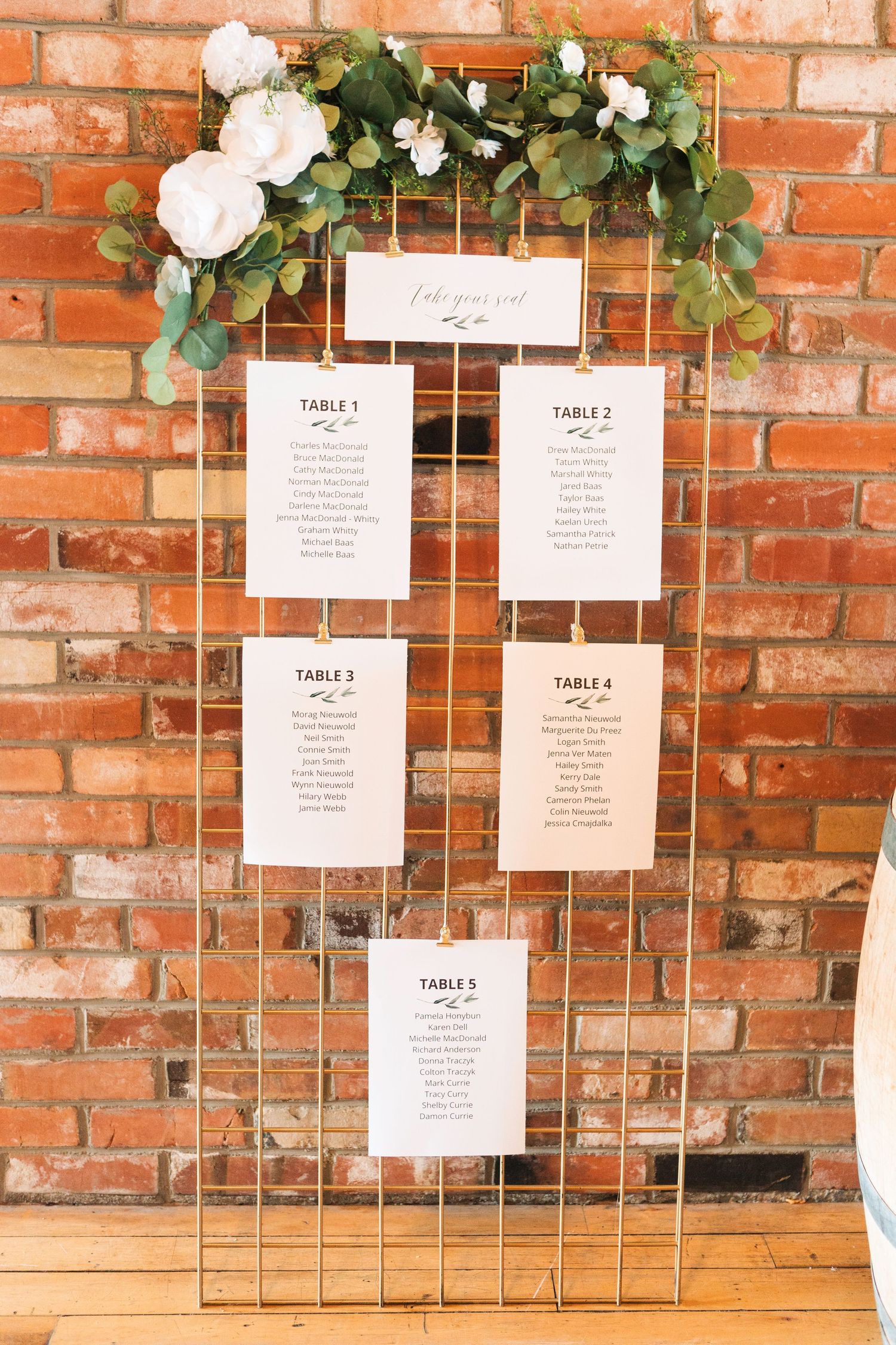 Wedding seating chart display with white cards mounted on metal grid against rustic brick wall with floral decorations.