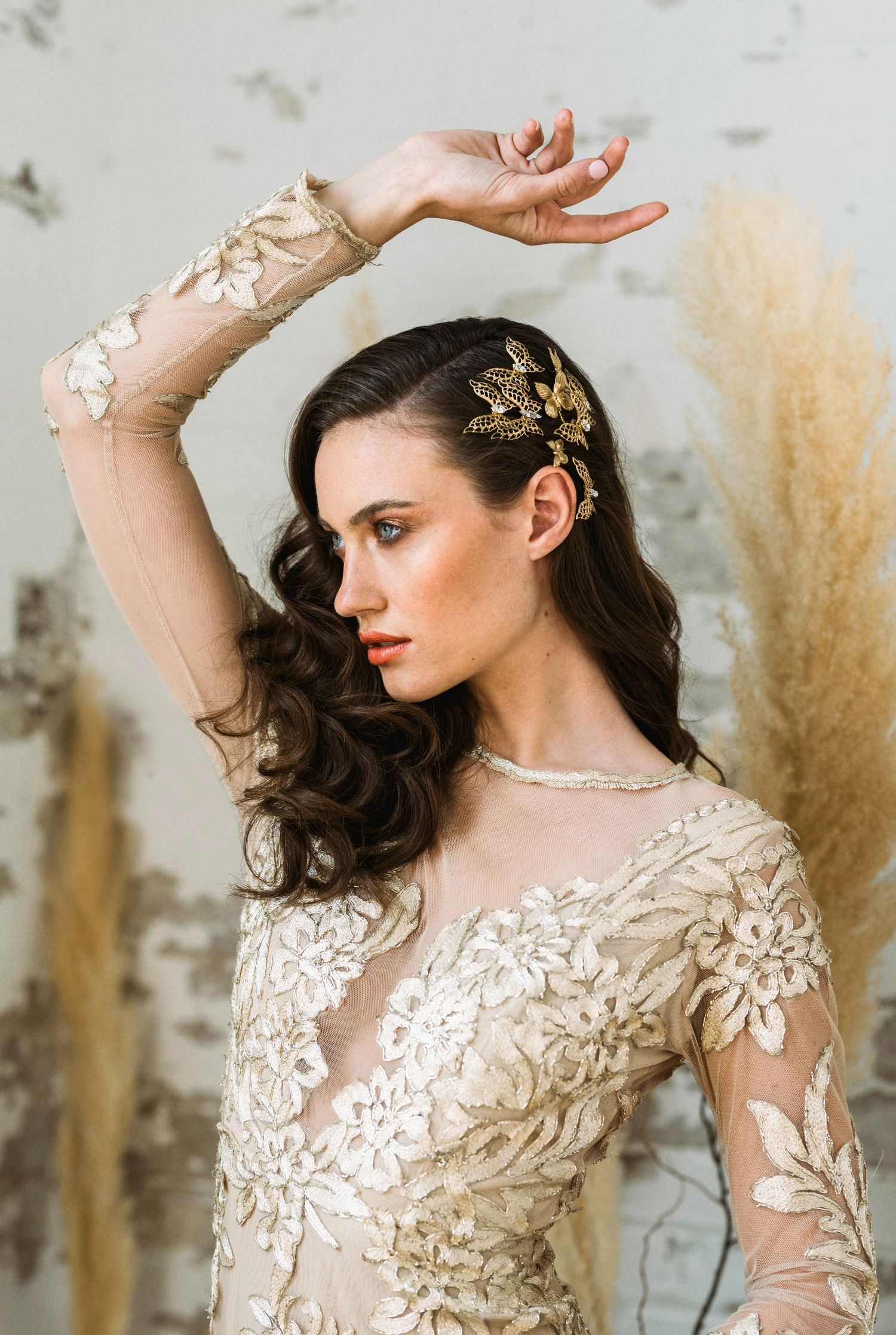 Model wearing intricate lace dress poses gracefully against neutral background with pampas grass.