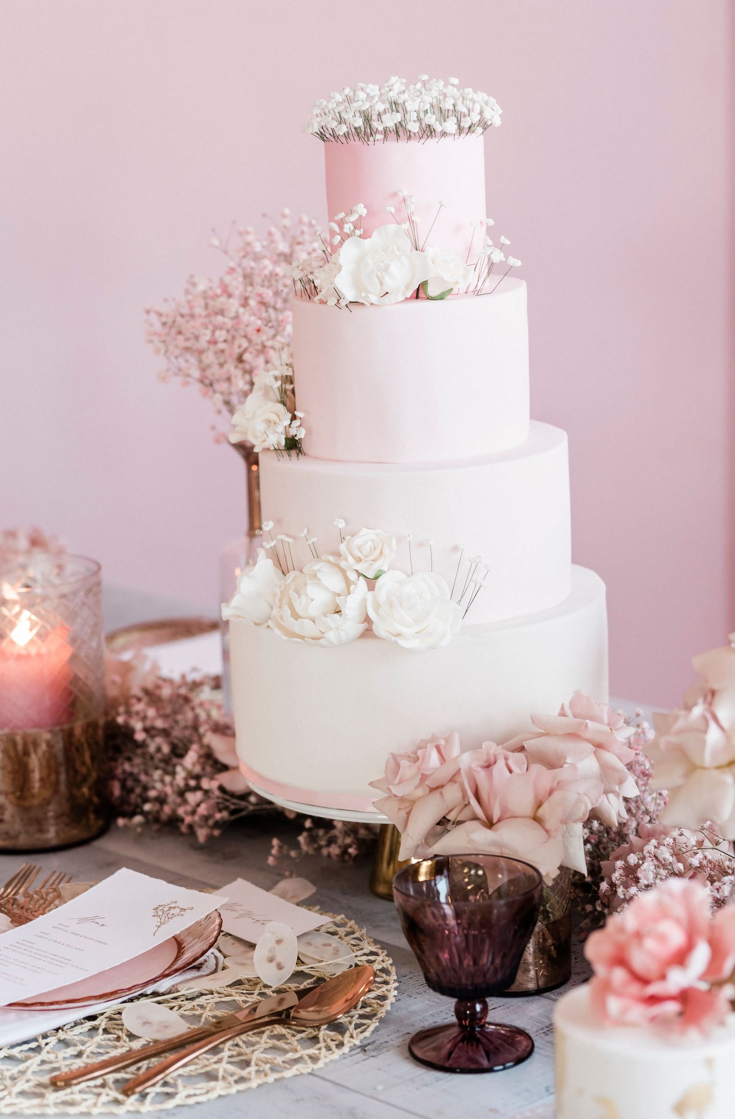 Elegant four-tier white wedding cake decorated with delicate flowers and baby's breath on a pink-toned table setting.