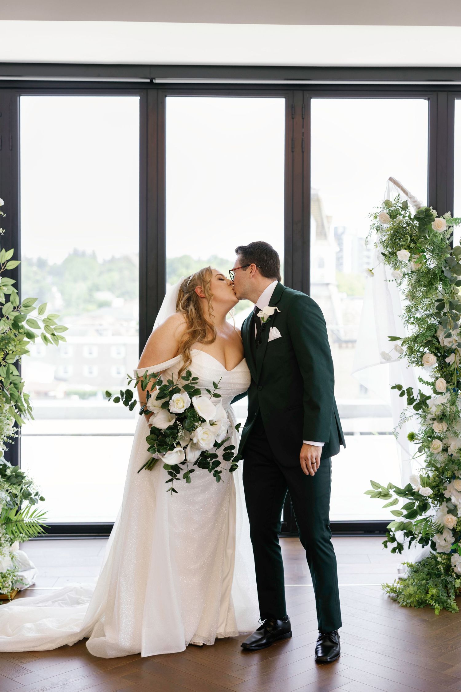 Newlyweds share a romantic kiss framed by floral arrangements and floor-to-ceiling windows.