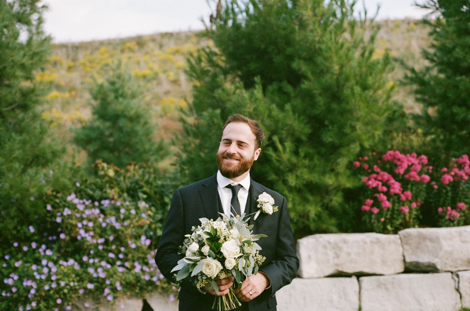 Smiling groom in dark suit holding white bouquet outdoors with colorful garden and stone wall backdrop.