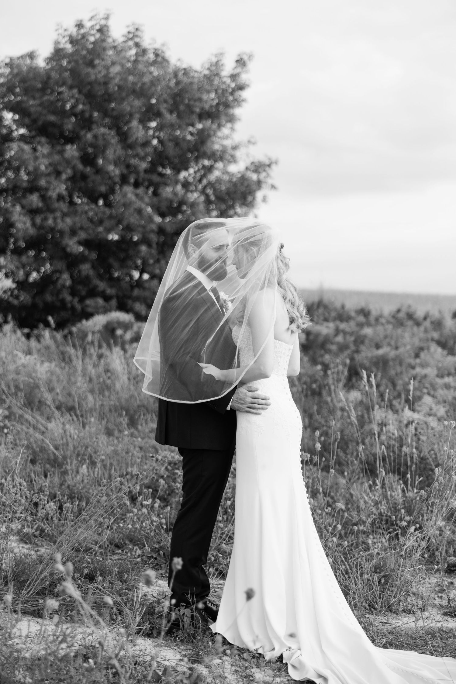 A romantic black and white wedding photo series showing a couple embracing outdoors in a natural setting with tall grass.
