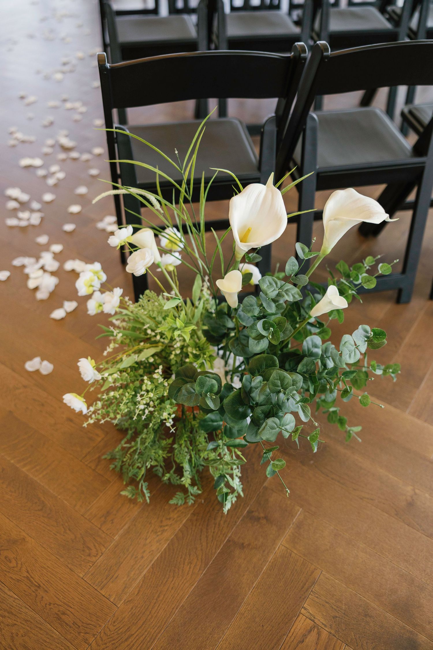 Beautiful floral arrangement with white calla lilies and greenery next to black ceremony chairs.