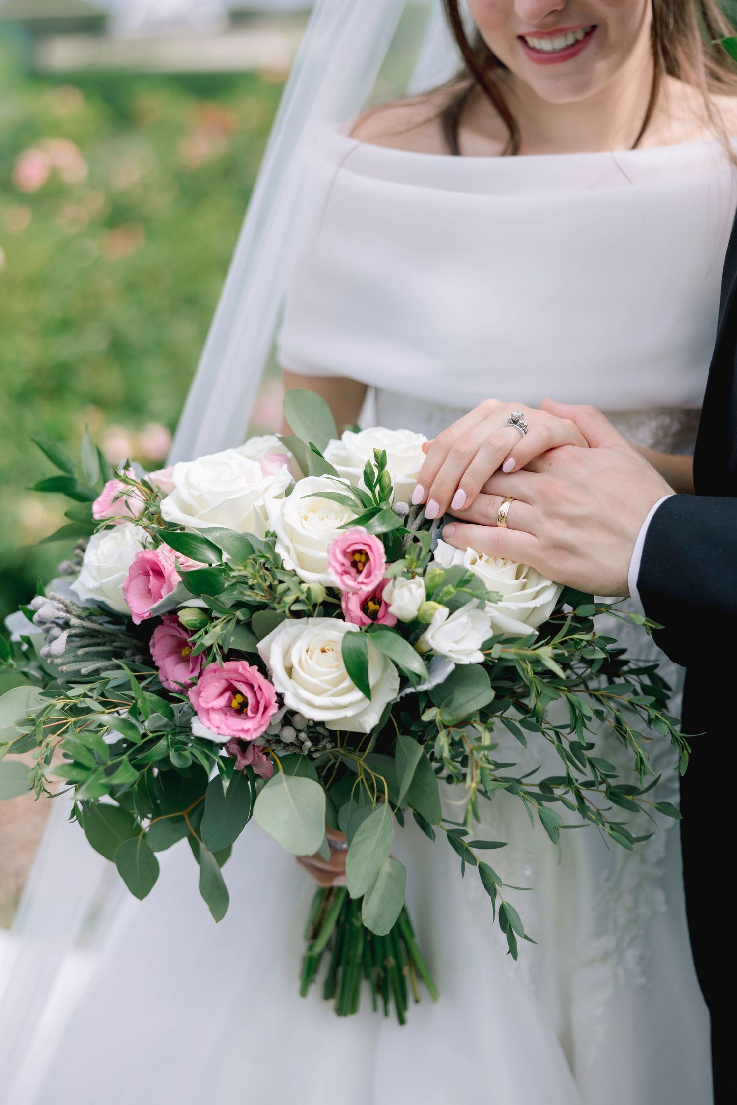 Bride and groom holding a beautiful wedding bouquet of white and pink roses with greenery, showing off their wedding rings.