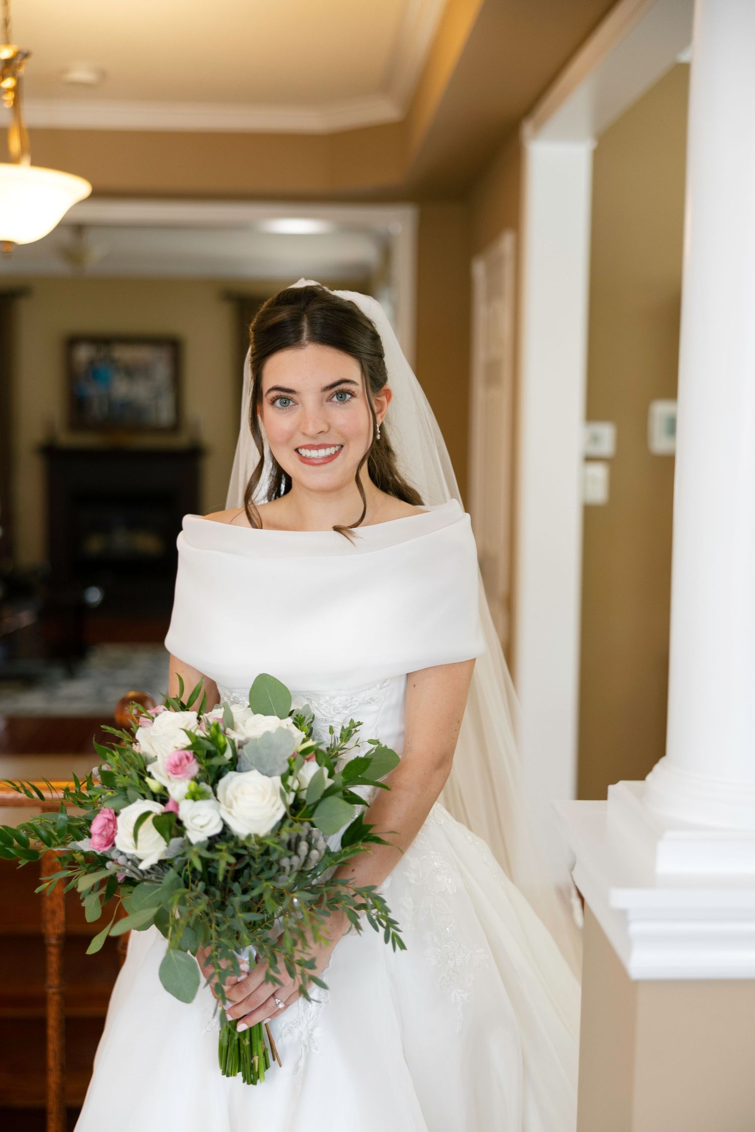 Smiling bride in white off-shoulder wedding gown holding lush bouquet of roses and greenery indoors.