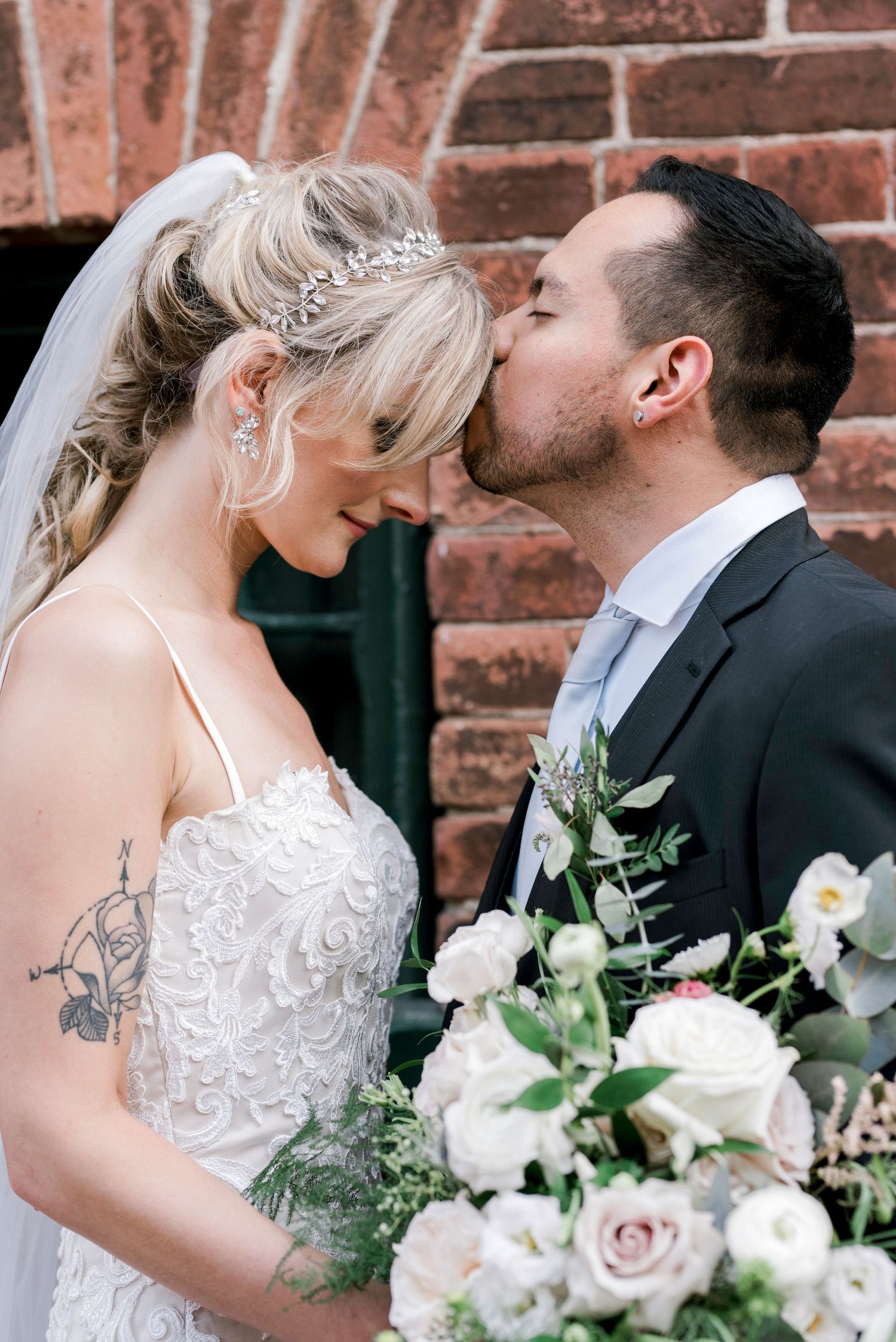Groom kissing bride on forehead, bride in lace dress with veil and hair vine, holding white floral bouquet outdoors.