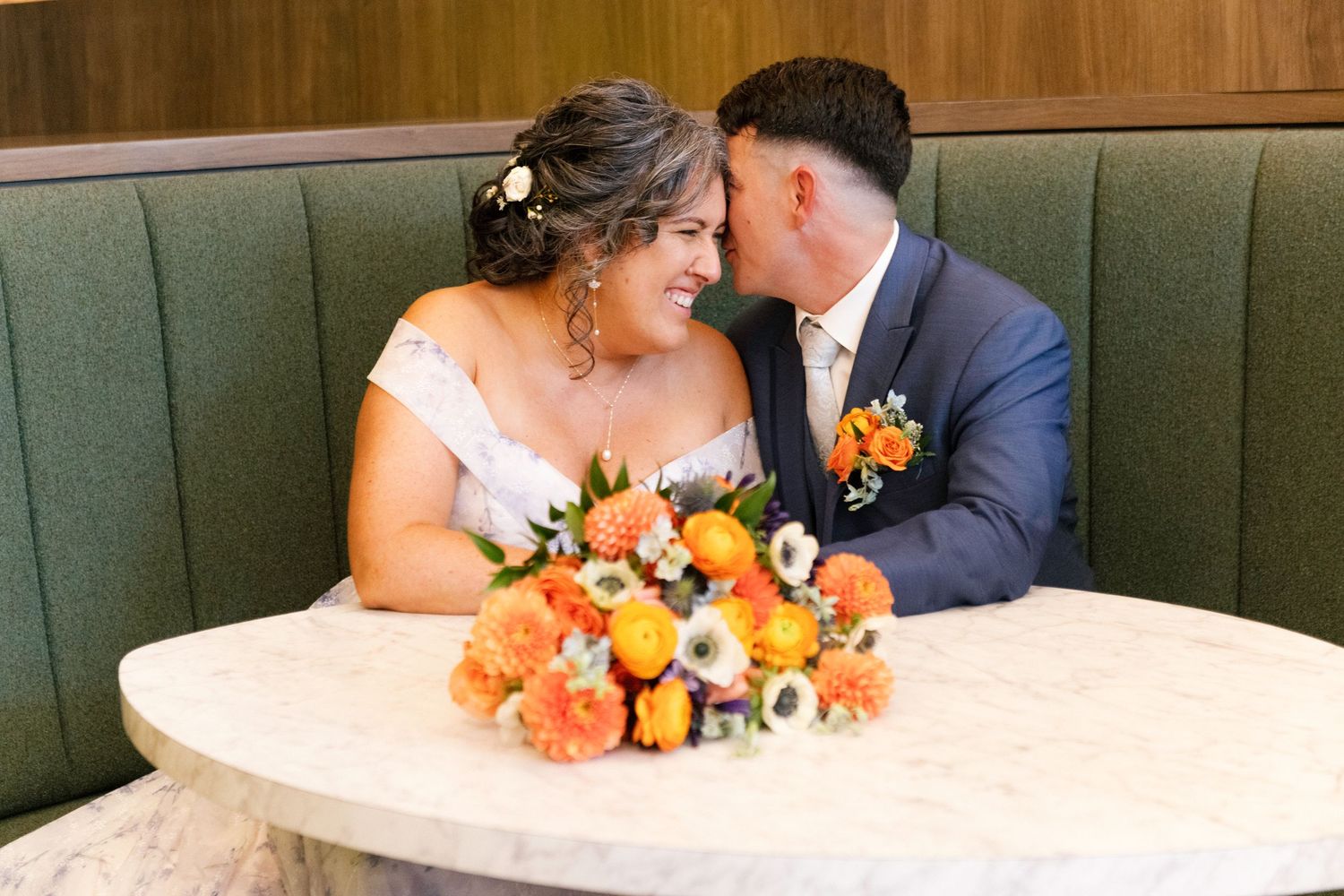 Bride and groom share a romantic moment in a green booth with orange floral bouquet on marble table.
