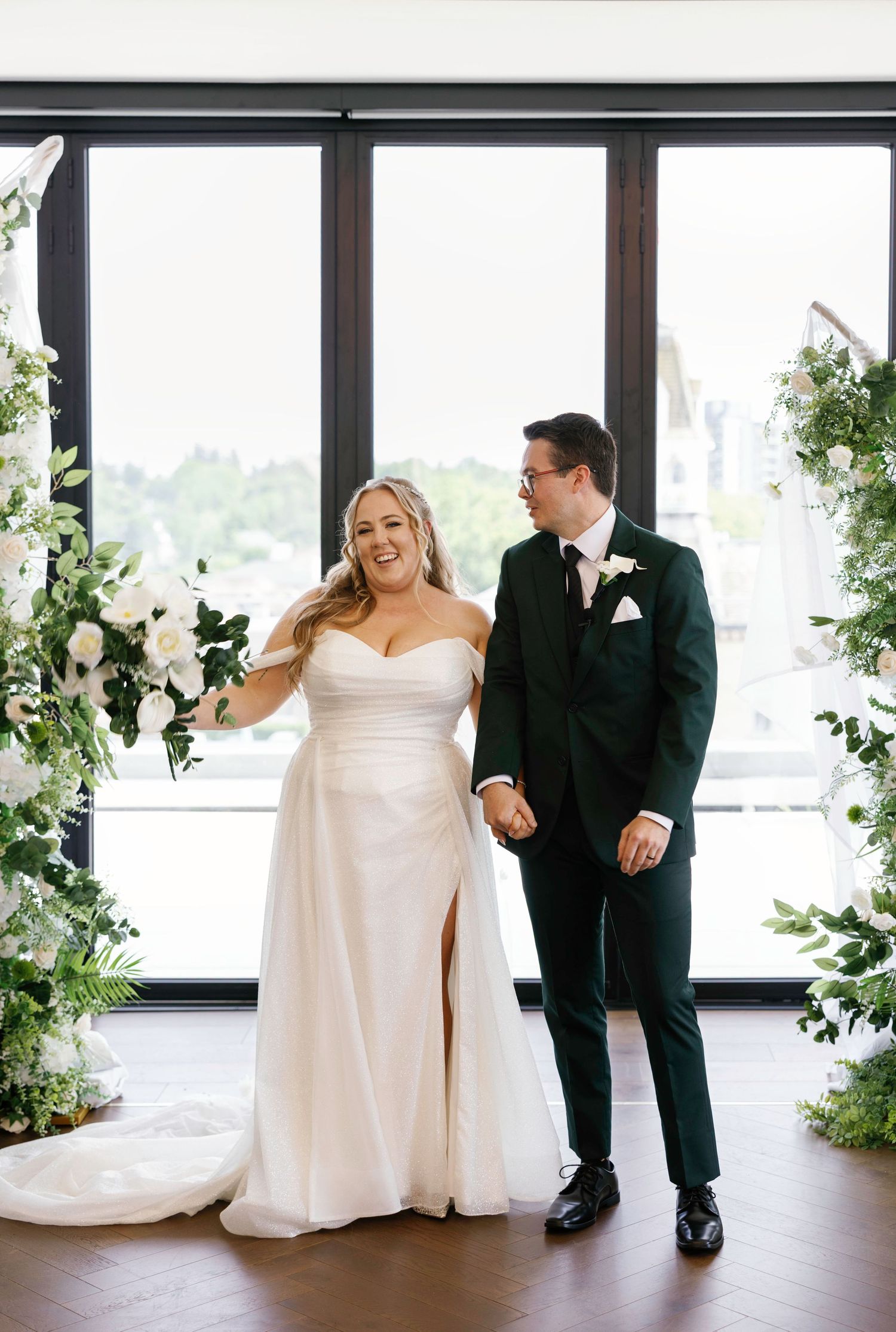 Wedding couple shares emotional first look in modern venue with floor-to-ceiling windows and white floral arrangements.
