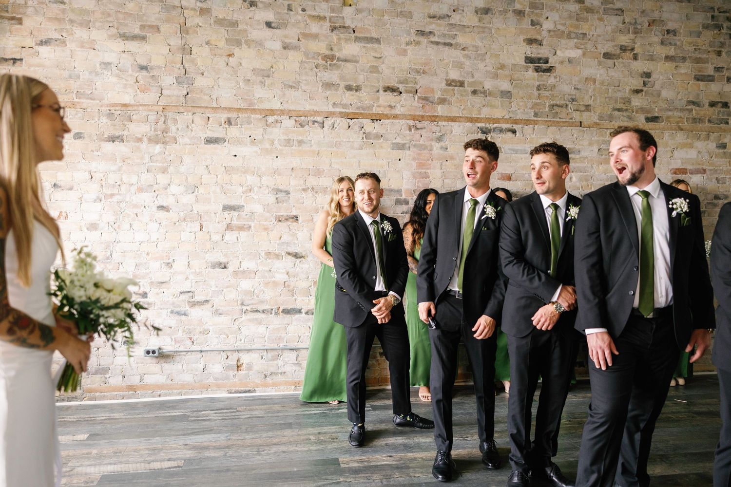 Groomsmen in dark suits and green ties pose together against a rustic brick wall at a wedding venue.