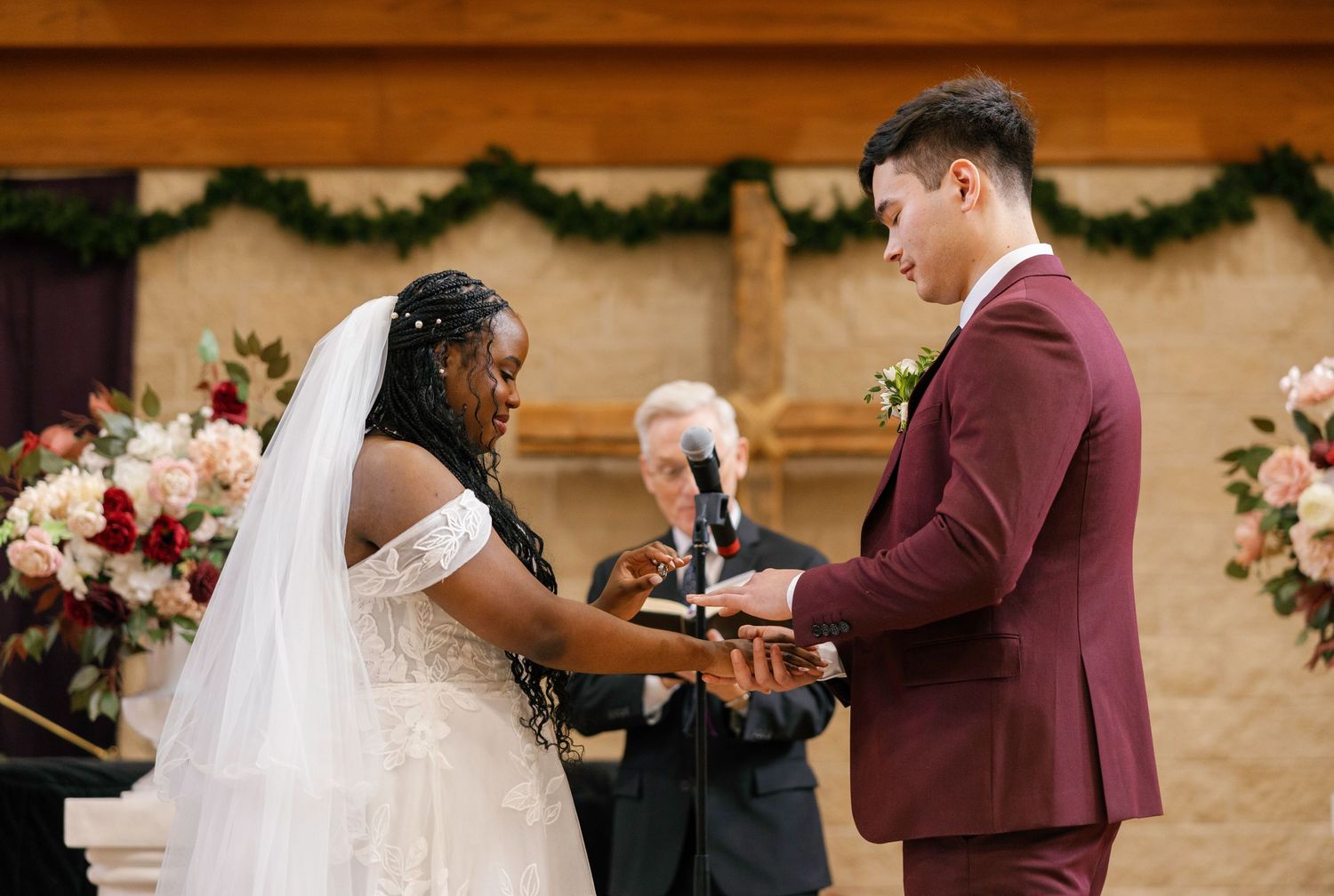 Bride and groom exchange rings at wedding altar, officiant behind them, floral arrangements and greenery in background.