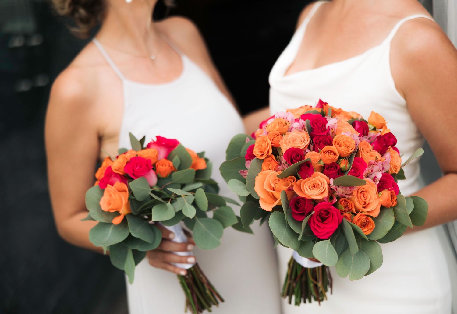 Two brides in white dresses holding vibrant orange and red rose wedding bouquets with eucalyptus greenery.