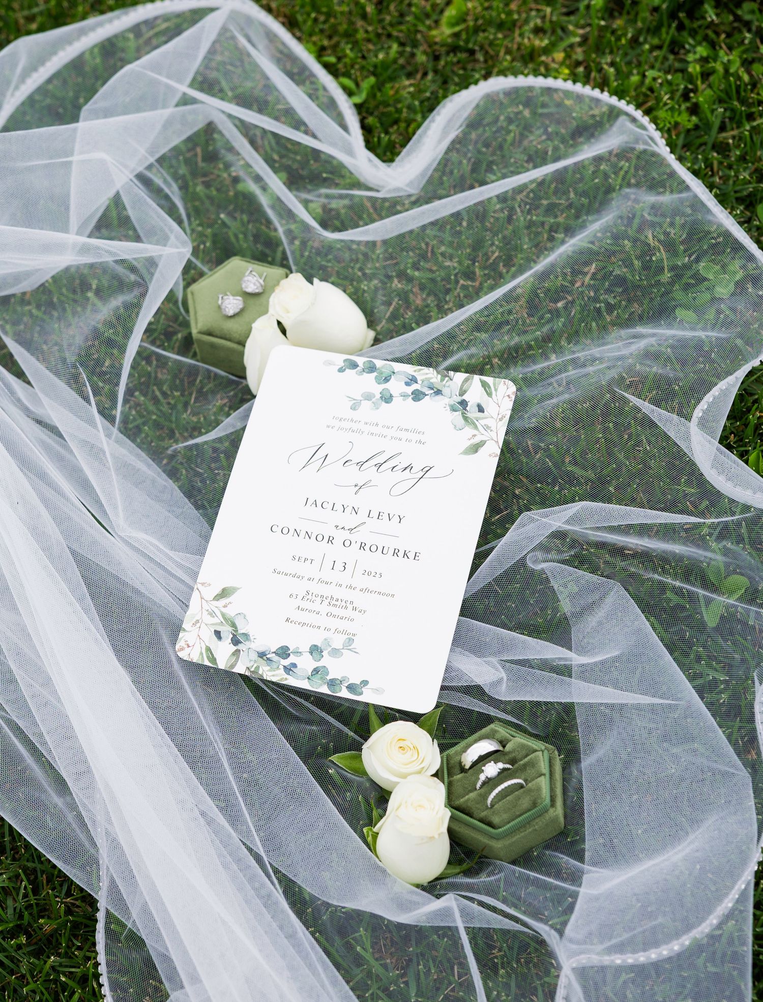 White tulle wedding veil draped elegantly over green grass with delicate floral wedding invitations and white roses.