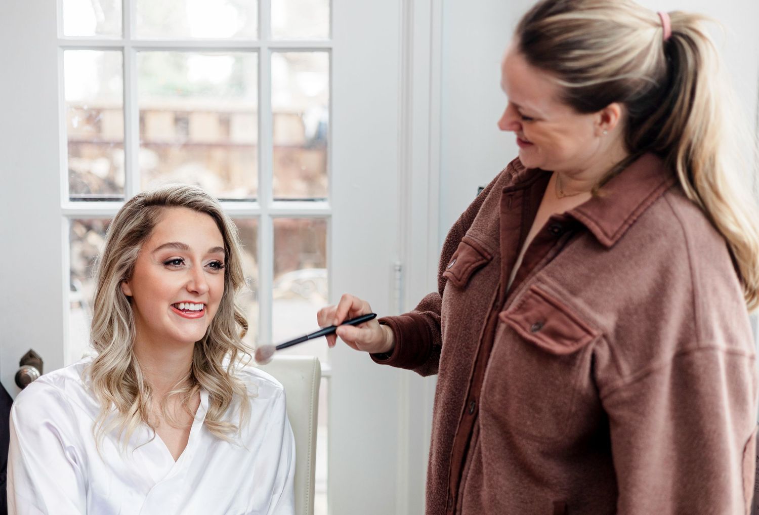 Beauty preparation scene with stylist working on client near bright window.