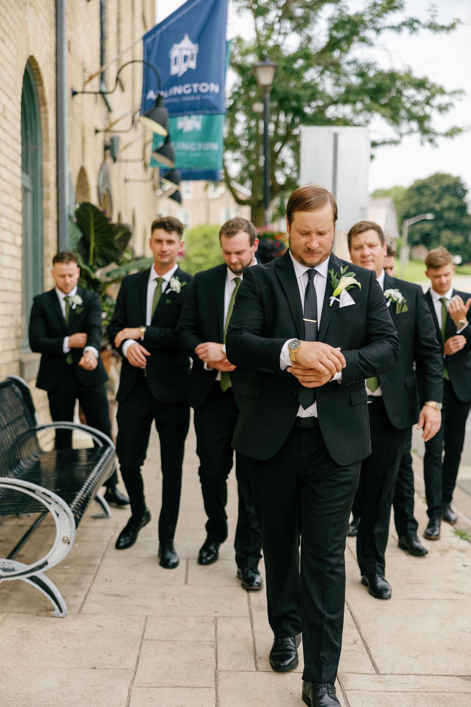 Groomsmen in black suits playfully dribble basketballs down a sidewalk outside a stone building.