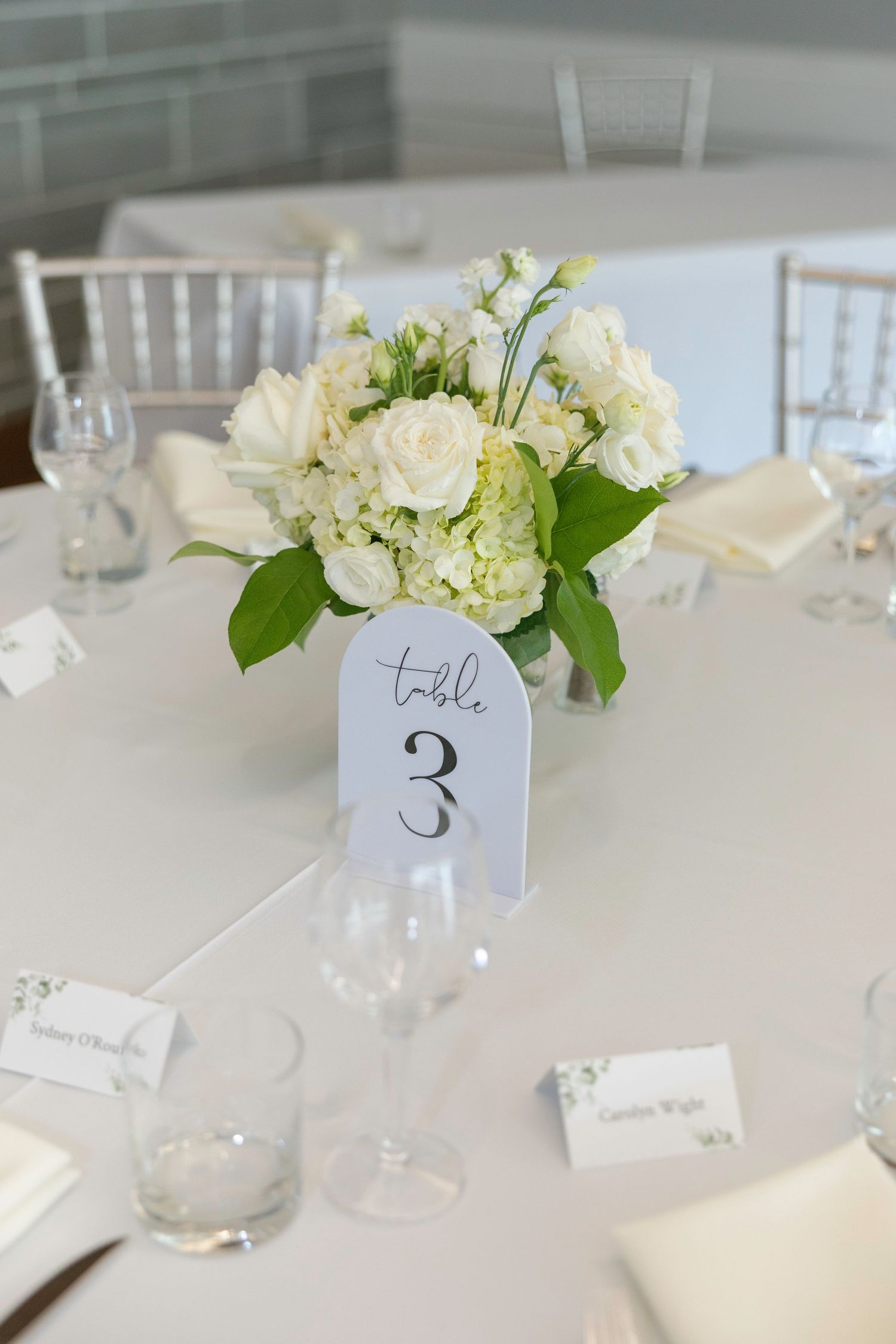 Elegant white floral centerpieces with table numbers decorate a wedding reception setup.