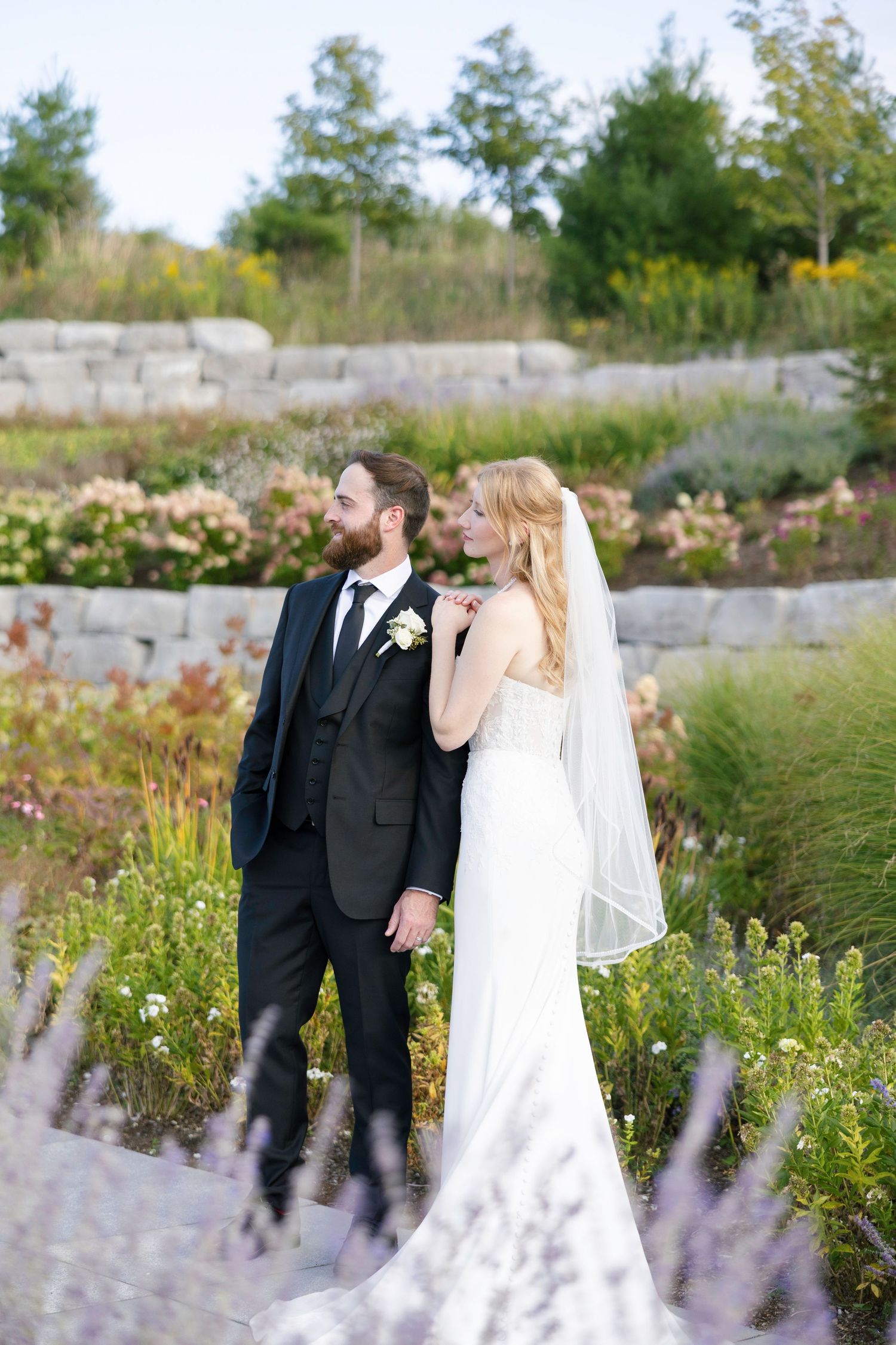 A romantic wedding couple poses in a garden filled with blooming lavender and wildflowers on a sunny day.