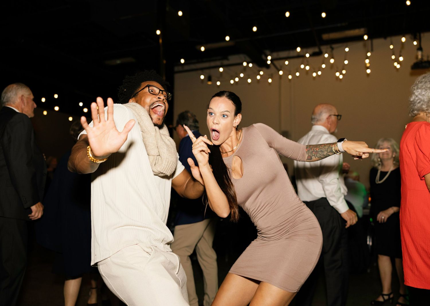 Two guests dance energetically at a wedding reception with string lights glowing in the background.