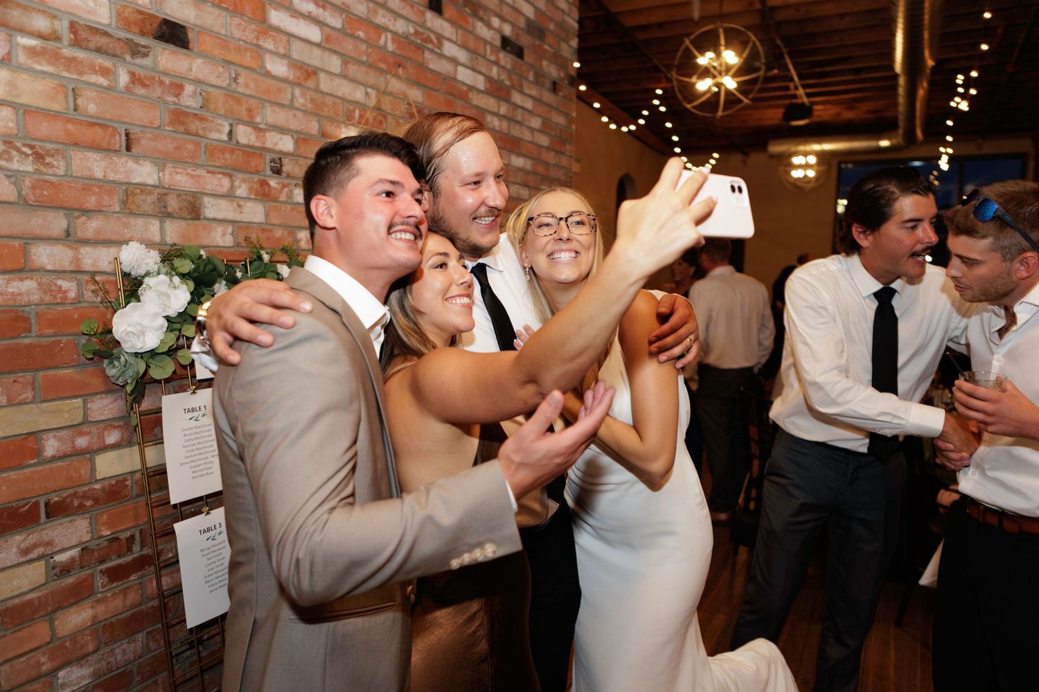 Wedding guests dance and celebrate on a dance floor against exposed brick walls with string lights overhead.