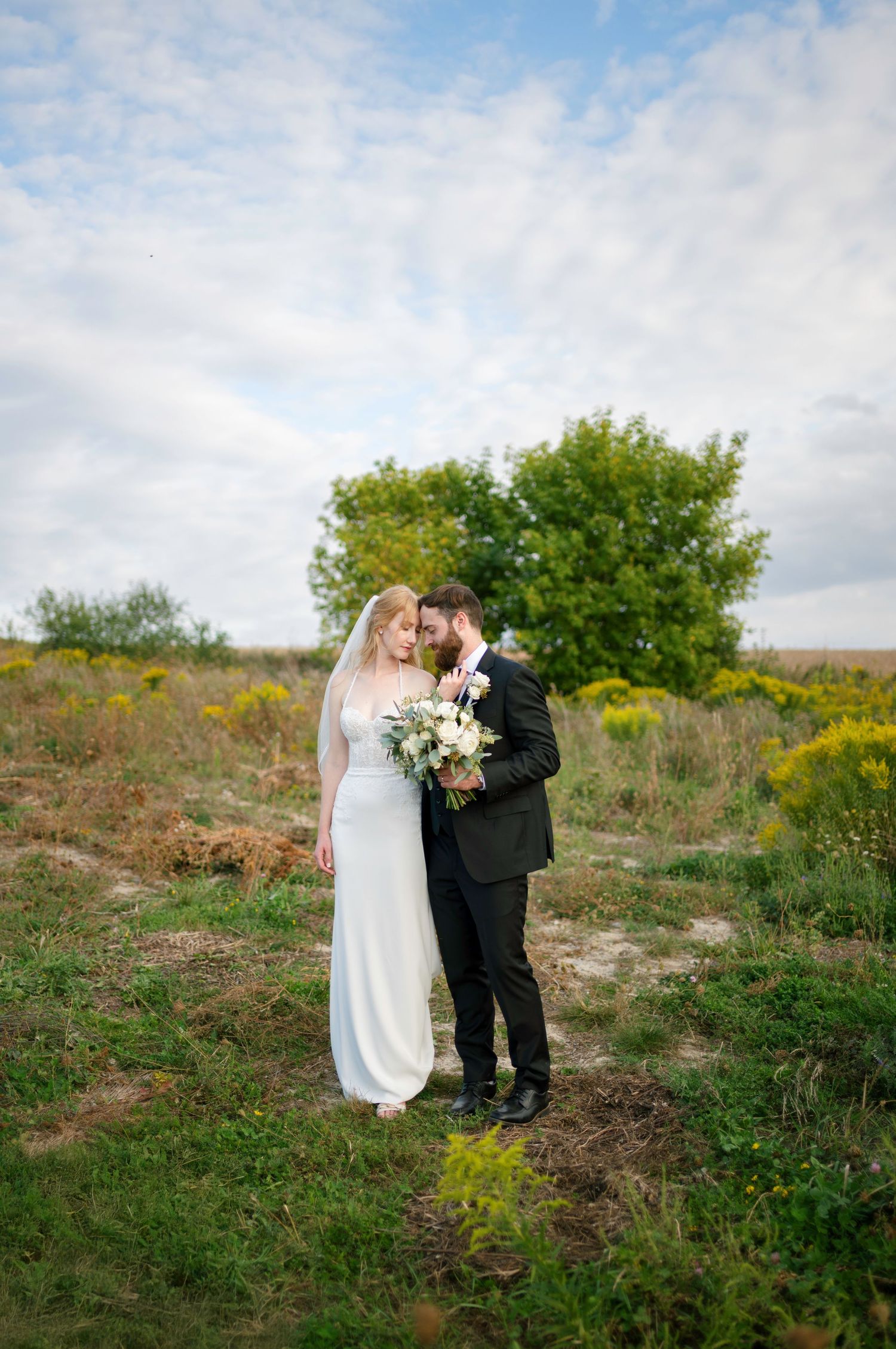 Newlyweds share an intimate moment on a hillside surrounded by greenery and wildflowers under a bright blue sky.