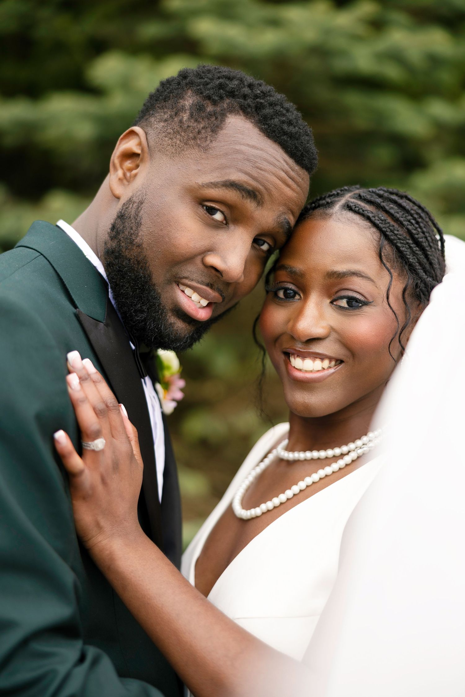 A joyful Black bride and groom pose cheek to cheek on their wedding day, she in white gown with pearls, he in a green suit.