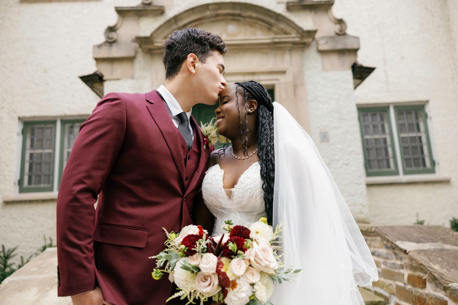 Interracial couple sharing a romantic kiss on their wedding day, groom in burgundy suit, bride in white gown holding floral bouquet.
