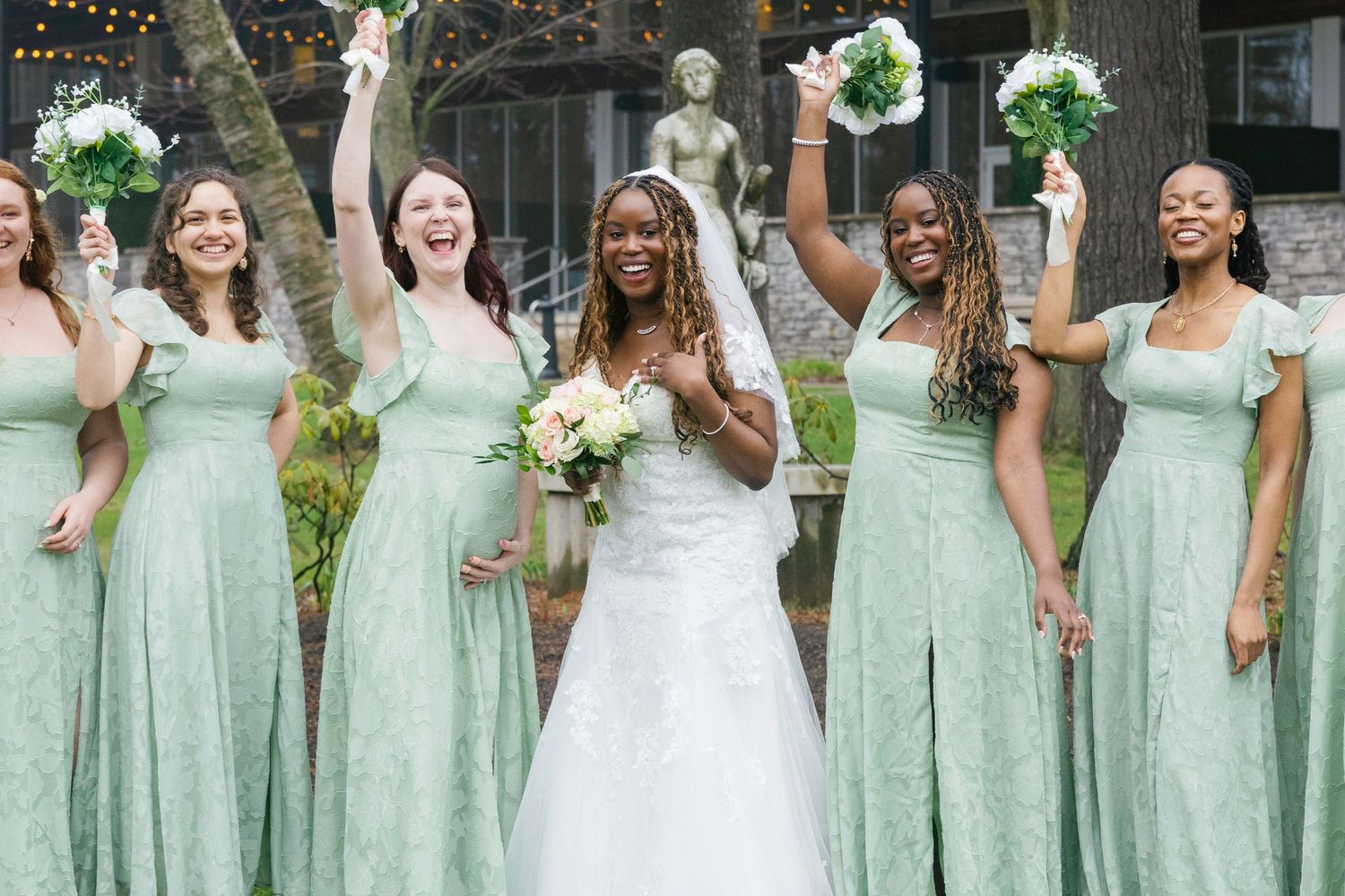 Bride in white gown poses with five bridesmaids in sage green dresses, all celebrating outdoors near a garden statue.