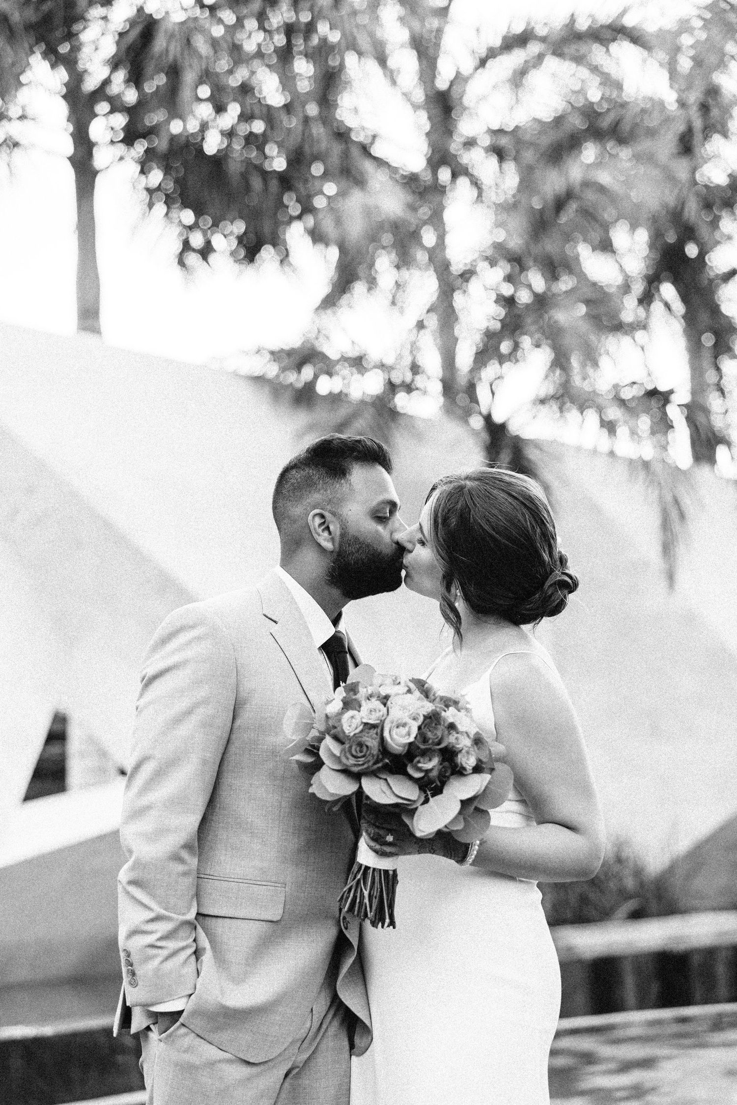 Black and white photo of a bride and groom sharing a kiss, bride holding a floral bouquet, palm trees in background.